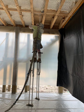 Worker on stilts installing spray foam insulation in a high ceiling.