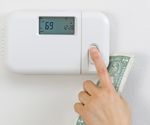 Closeup of a hand holding US dollars and adjusting a thermostat.